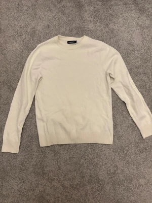 Vit stickad tröja från J.Lindeberg - Snygg vit stickad tröja från J.Lindeberg i storlek S. Tröjan har ett diskret strukturerat mönster och rund hals. Långärmad med ribbade muddar vid ärmslut och nederkant. Perfekt för dig som gillar stilrena och klassiska plagg.