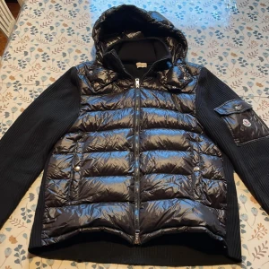 Svart pufferjacka från Moncler - Snygg svart pufferjacka från Moncler med glansig dunfylld front, stickade ärmar och huva. Jackan har dragkedja, fickor med tryckknappar och Moncler-logga på ärmen. Perfekt för dig som vill ha en stilren och exklusiv look under kyliga dagar.