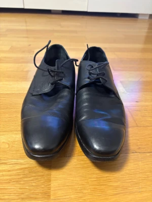 Svarta skinnskor från Tiger of Sweden - Snygga svarta skinnskor från Tiger of Sweden med klassisk rund tå och snörning. Skorna har låg klack och en stilren, minimalistisk design som passar perfekt till kostym eller dressade outfits. Tillverkade i mjukt skinn för en exklusiv känslan. Riktigt läder, storlek 45