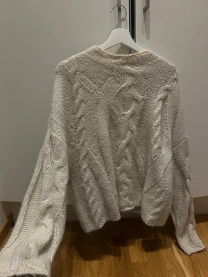 Beige stickad tröja med flätmönster - Säljer en mysig beige stickad tröja med grova flätor och kabelstickat mönster. Tröjan har lång ärm och en loose passform som ger en chill vibe. Perfekt för att hålla sig varm under kalla dagar och passar till jeans eller kjol.