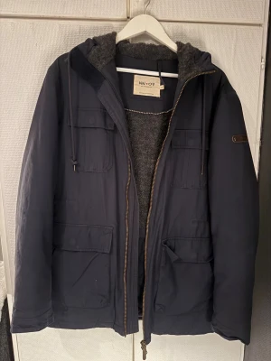 Mörkblå parkajacka från NN07 - Parkajacka från NN07 i mörkblå färg med huva och grått foder. Jackan har flera stora fickor framtill, dragkedja och snörning vid huvan. Perfekt för kalla dagar med sin rejäla och stilrena design.