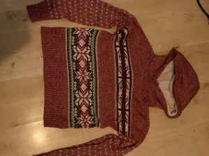Röd hoodie med vintermönster från Yet Again - Säljer en röd hoodie från Yet Again i storlek L med mysig fuskpäls i luvan. Tröjan har ett vintrigt mönster i vitt och grönt över bröstet och armarna, samt långa ärmar och ribbade muddar. Perfekt för kalla dagar och chill hemma. Storlek L men sitter som M