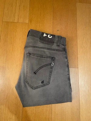 Gråa Dondup - Säljer ett par grå jeans med skinny passform och coola slitningar på både fram- och bakfickor. Jeansen har klassisk femficksdesign, svarta sömmar och en vit detalj på bälteshällan. Perfekt för dig som gillar en edgy stil.