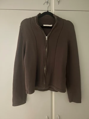 Brun kofta från B-Young - Brun kofta från B-Young i storlek M. Koftan har ribbad krage, långa ärmar och dragkedja framtill. Snygga hålstickade detaljer längs axlar och ärmar.💞