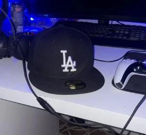 Svart LA 59FIFTY keps från New Era - Säljer en svart New Era 59FIFTY keps med LA-logga i vitt framtill. Klassisk flat brim och broderad New Era-logga på sidan. Kepsen är tillverkad i polyester och har en stilren look som passar till streetwear. Perfekt för dig som gillar sportiga vibes och snygga accessoarer.