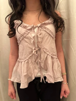 Ljusrosa blus med volanger och snörning - Supersöt ljusrosa blus (ljuset ändrar färg på blusen) med volanger och snörning framtill. Blusen är ärmlös och har flera dekorativa volangdetaljer som ger en romantisk vibe. Tillverkad i ett lätt och luftigt material som känns skönt mot huden. Perfekt för dig som gillar en feminin och trendig stil.