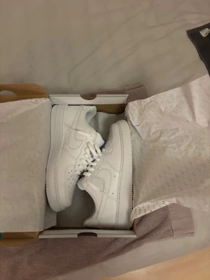 Nike Air Force 1 '07 vita sneakers - Nike Air Force 1 '07 sneakers i helvit färg med klassisk låg siluett. Skorna har snygga detaljer som perforerad tå, snörning och rund tå. Tillverkade i skinn med platt sula och ikonisk Swoosh på sidan. Perfekt för dig som gillar clean och tidlös streetstil.