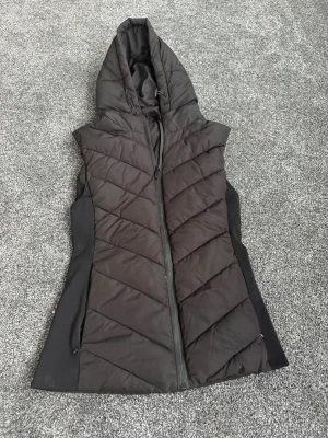 Svart quiltad dunväst med huva - Säljer en svart quiltad dunväst med stor huva och dragkedja framtill. Västen har diagonala sömmar för en snygg look och ribbade sidopaneler som ger en skön passform. Perfekt för lager på lager under kyliga dagar. Dåliga bilder, färgen är helt svart