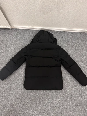 Canada goose Macmillian svart badge - Helt ny aldrig använd Svart pufferjacka från Canada Goose med huva och quiltad design. Jackan är tillverkad i polyester och dun, har långa ärmar och en stilren look. Perfekt för kalla dagar och har innerficka med dragkedja. Märke och storlek syns tydligt på insidan.