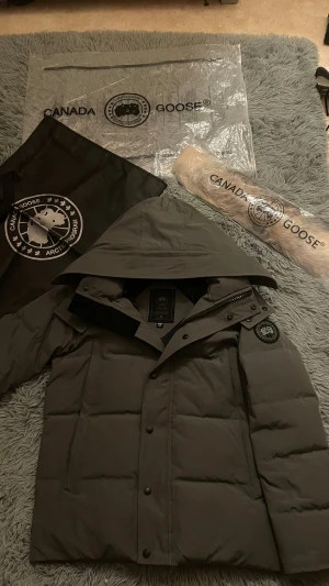 Grå Canada Goose Wyndham  - Säljer en grå dunjacka från Canada Goose med stor huva och avtagbar beige päls. Jackan har svarta detaljer, dragkedja och knappar framtill samt den klassiska Canada Goose Arctic Program-patchen på ärmen. Perfekt för kalla vinterdagar och riktigt snygg stil.