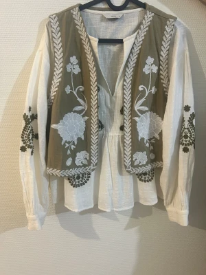 Broderad overshirt från Pigalle - Säljer en snygg overshirt från Pigalle i beige och vitt med gröna och vita broderade blommor. Jackan har långärmade ärmar, v-ringad hals och coola detaljer på både fram- och baksida. Materialet är lätt och känns fräscht, perfekt för vår eller sommar.