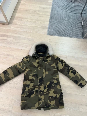 Camouflage parkas från Frilivin med päls - Parkas från Frilivin i storlek L med grön, brun och svart camouflage-mönster. Jackan har en fluffig vit fuskpäls runt huvan, dragkedja och knappar framtill samt stora fickor. Perfekt för kalla dagar och har en cool streetstil.