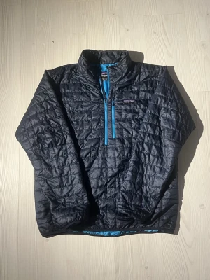 Svart pufferjacka Patagonia XL - Quiltad svart pufferjacka från Patagonia i storlek XL. Jackan har blå detaljer, halv dragkedja framtill och en bröstficka med dragkedja. Lätt och smidig modell med hög krage, perfekt för kyliga dagar. I perfekt skick