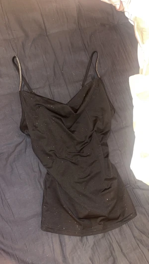 Svart glittrig linnetopp med smala band - Svart linnetopp med glittriga detaljer och smala axelband som har glitter detaljer pp. Toppen har en snygg draperad v-ringning och är figurnära. Perfekt för dig som gillar att sticka ut med lite extra glitter och en cool siluett. NYÅRS TRÖJA💓