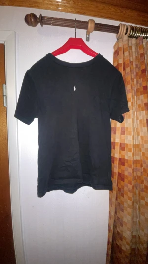 Svart t-shirt från Ralph Lauren - Svart t-shirt från Ralph Lauren med klassisk rund hals och korta ärmar. Diskret vit logga broderad på bröstet. Tillverkad i mjuk bomull som känns skön mot huden. Enkel och stilren design som funkar till det mesta.