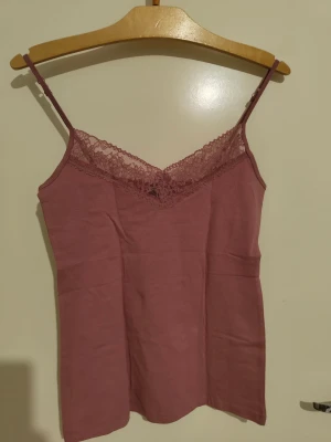 Rosa linnetopp med spetsdetalj - Supersöt rosa linnetopp med tunna axelband och v-ringad spetskant framtill.Man kan ha den under nån tröja med spetsen som syns, ger lite downtown girl vibe, jättegulligt. Toppen har en mjuk och skön känsla, perfekt för varma dagar. Spetsen ger en extra touch av stil och gör toppen lite mer unik. Enkel att matcha med jeans eller kjol. 