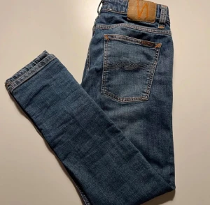 Blå jeans från Nudie Jeans - Snygga blå jeans från Nudie Jeans med klassisk femficksdesign och bruna sömmar. Modellen har en rak passform med något smalare ben och normal midja. Jeansen är tillverkade i slitstarkt denim och har Nudie-loggan på bakfickan. Knappt använt, har testat och priset är sjukt lågt så hugg till…Vid minsta fundering kontakta mig(svara extremt snabbt)😉🙏