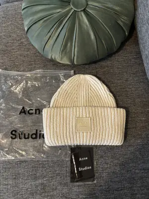 Säljer en snygg vit stickad mössa från Acne Studios med klassisk ribbad design och den ikoniska ansiktsloggan framtill. Mössan är tillverkad i 100% ull och har en bred uppvikt kant. Perfekt för att hålla värmen och samtidigt se clean ut.