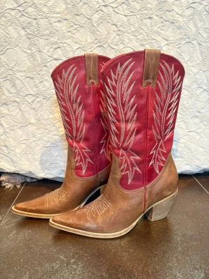NYA Bruna och röda cowboy boots i äkta skinn stl.37 - Snygga cowboy boots i brunt och rött skinn med dekorativa broderier i vitt. Skorna har spetsig tå, låg klack och klassisk western-design. Perfekta för dig som vill sticka ut med en cool och tidlös look. Aldrig använda. Innermått 23 cm