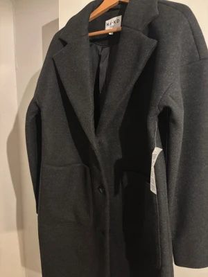 Mörkgrå kappa från NA-KD, strl 34 - Snygg mörkgrå kappa från NA-KD i klassisk rak modell med stora fickor och två knappar framtill. Jackan har bred krage och är helfodrad, perfekt för dig som gillar stilrena och enkla plagg. Materialet är mjukt och känns lyxigt.