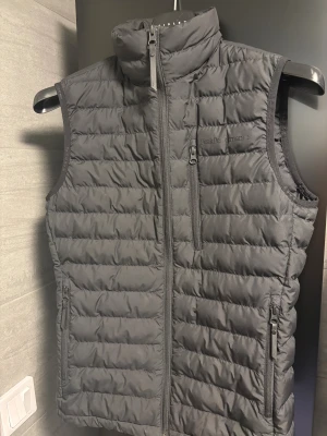 Svart dunväst från Peak Performance - Svart dunväst från Peak Performance med quiltad design och hög krage. Västen har tre praktiska fickor med dragkedja och diskret logga på bröstet. Perfekt för lager-på-lager och outdoor-aktiviteter. Snygg, stilren och funktionell.