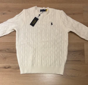 Vit kabelstickad tröja Polo Ralph Lauren - Snygg vit kabelstickad tröja från Polo Ralph Lauren med klassisk broderad logga på bröstet. Tröjan har rund hals, ribbade muddar och är tillverkad i mjuk bomull. Perfekt för dig som gillar stilrena och tidlösa plagg.