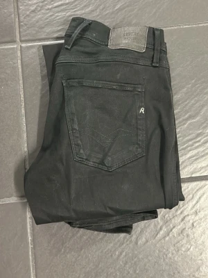 Svarta Replay Anbass jeans W29 L32 - Svarta jeans från Replay, modell Anbass. Klassisk femficksdesign med Replay-logga på fickan och bakre läderpatch. Slimmad passform och tillverkade i mjukt bomullstyg med stretch för extra komfort. Perfekta för dig som gillar stilrena svarta jeans.