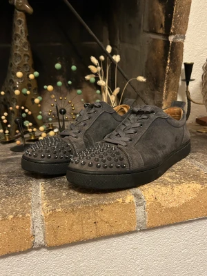 Christian Louboutin grå mocka sneakers - Säljer ett par gråa sneakers från Christian Louboutin i mocka med coola svarta nitar på tån och klassisk röd sula. Skorna har snörning och en rund tå, samt en platt sula. Insidan är beige och de har en riktigt edgy vibe som sticker ut.