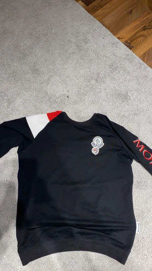 Svart långärmad Moncler tröja - Svart långärmad tröja från Moncler med broderad logga på bröstet och stor röd Moncler-text på ena ärmen. Ena axeln har en cool detalj i vitt och rött. Tröjan är i mjuk bomull och har en normal passform. Perfekt för dig som gillar stilrena märkesplagg.