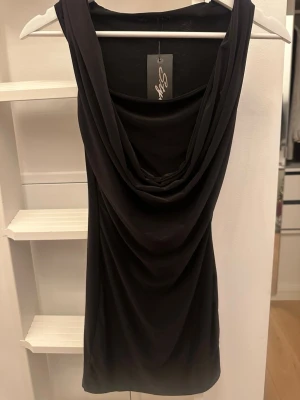 Svart draperad ärmlös klänning  - Svart ärmlös tajt klänning med snygg draperad front och djup halsringning. Toppens tyg faller mjukt och ger en cool, elegant vibe. Perfekt att styla med kjol för en trendig look. Materialet är lite mot träningsmaterial, mjukt och skönt mot huden. 