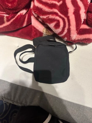 Svart axelväska från Adidas - Säljer en svart axelväska från Adidas med justerbar rem och dragkedja. Väskan har en enkel, stilren design med Adidas-loggan i vitt framtill. Perfekt storlek för mobil, plånbok och andra småsaker. Tillverkad i slitstark polyester.