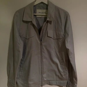 Grå vindjacka från These Glory Days - Snygg grå overshirt jacka från These Glory Days med klassisk krage, två bröstfickor med lock och sidofickor. Jackan har dragkedja framtill och knappar vid ärmslut. 