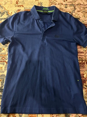 Blå pikétröja från Hugo Boss - Säljer en stilren blå pikétröja från Hugo Boss med klassisk krage och knappar framtill. Tröjan har broderad Boss-logga på bröstet och är tillverkad i bomull med fuktavledande egenskaper. Perfekt för dig som gillar en clean och sportig look.