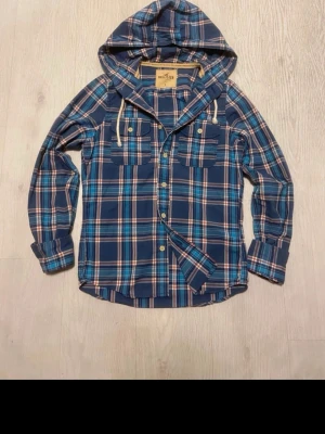 Blårutig overshirt från Hollister - Snygg blårutig overshirt från Hollister med huva och snörning. Jackan har två bröstfickor med knappar och långa ärmar. Perfekt för dig som gillar avslappnad stil och vill ha något som sticker ut med sitt rutiga mönster och coola detaljer.