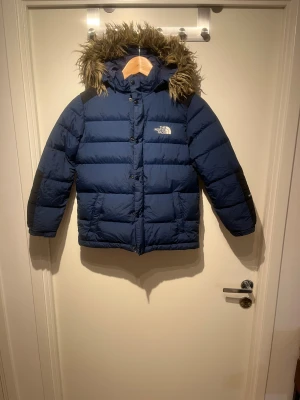  The North Face (barn storlek) - Blå North Face dunjacka barn, storlek 130 (passar ca 7–9 år). HyVent-material, svarta ärmdetaljer och avtagbar huva med fejkpäls. Varm och perfekt för vinter. Pris kan diskuteras – skicka gärna bud.