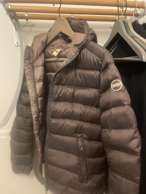 Brun Colmar dunjacka med huva - Säljer en brun dunjacka från Colmar med huva och två sidofickor med dragkedja. Jackan har ett quiltat mönster och Colmar-logga på ärmen. Perfekt för kalla dagar och har en snygg, klassisk look med lång ärm och normal passform. (OBS drag kedjan e sönder men går att fixa)
