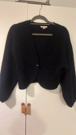 Svart stickad kofta från Gina Tricot - Mysig svart stickad kofta från Gina Tricot med v-ringad hals och två knappar framtill. Koftan har långärmade, lite loose ärmar och är tillverkad i ett fluffigt stickat material som känns skönt mot huden. Perfekt för att addera lite extra värme till din outfit.