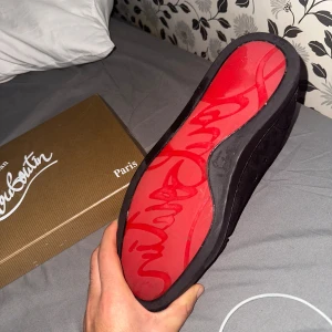Christian Louboutin svarta fin skor med signatur - Svarta sneakers från Christian Louboutin med lyxig röd sula och signatur på undersidan. Skorna har snörning, rund tå och är tillverkade i mocka med diskret broderad logga på hälen. Insidan är ljus och de levereras med originalkartong och dustbag.