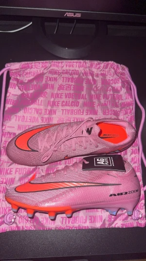 Nike Mercurial rosa fotbollsskor - Nike Mercurial fotbollsskor i en snygg rosa färg med orange detaljer och svart logga. Skorna har snörning, platt sula med dobbar och är tillverkade i syntetmaterial för bästa grepp på planen. Perfekta för dig som vill sticka ut på fotbollsplanen.
