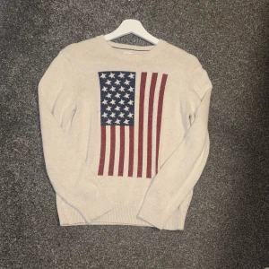 Beige stickad tröja GAP Kids USA-flagga - Snygg beige stickad tröja från GAP Kids med stort USA-flagga tryck på framsidan. Tröjan har rund hals, ribbade muddar och långa ärmar. Perfekt för dig som gillar klassisk stil med en cool twist. Storlek 164