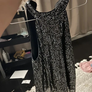 Glittrig svart paljettblus  - Supersnygg svart klänning med massor av silvriga paljetter och hög hals. Ärmlös modell med rak passform som ger en cool och festlig look. Perfekt för dig som vill sticka ut med lite extra bling. Klänningen är kort och har en lätt A-linjeform.