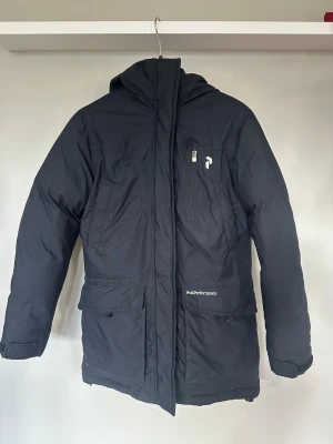 Peak Performance XS vinterjacka parkajacka - Svart parkajacka från Peak Performance i storlek XS. Jackan har huva, flera fickor med dragkedja och logga på bröstet och huvan. Materialet är slitstark polyester med dunfoder och meshdetaljer på insidan. Trasig mesh på innerfickorna, i övrigt i fint skick. Därav lågt pris. 