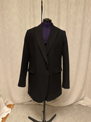 Svart kavaj från JJXX - Stilren oversized svart kavaj från JJXX med klassisk passform och enkla knappar framtill. Kavajen har två fickor med lock och en bröstficka, samt slits bak. Fodret är i en kontrasterande beige nyans. Perfekt för dig som vill ha en tidlös och snygg look. Ny pris ligger runt 699-799kr