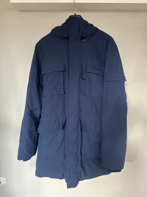 Carhartt herr strl M vinterjacka med huva parkas - Säljer en mörkblå parkas från Carhartt med huva strl M. Flera stora fickor framtill. Jackan har snygga detaljer som metallknappar med logga och dragsko vid halsen. Lite slitage på knappar. Använt fint skick. 