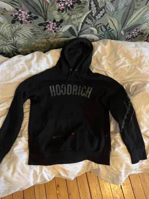 Svart HOODRICH hoodie med tryck - Svart hoodie från HOODRICH med stor logga på bröstet och tryck på ena ärmen. Har känguruficka och huva med snören Snygga detaljer och text även på ryggen. Perfekt för dig som gillar att se snygg ut