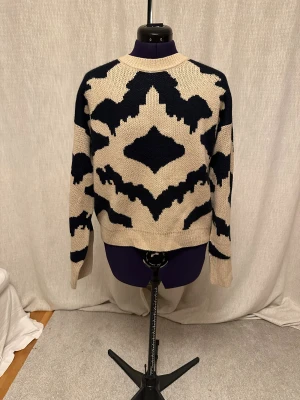 Beige och svart stickad tröja - Snygg stickad tröja i beige och svart med ett grafiskt mönster som täcker hela plagget. Tröjan har rund hals, ribbade muddar och långärmade ärmar. Perfekt för dig som gillar statement-plagg med unik design och skön passform.