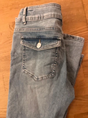 Bootcut ljusblå jeans från Kappahl - Snygga ljusblå bootcut jeans med dubbla knappar i midjan och klassiska fickor, inklusive en ficka med lock och knapp bak. Jeansen har en rak passform över låren och lätt utsvängda ben. Perfekta för dig som gillar retrovibbar och bekväm stil.