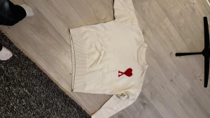 Vit stickad tröja med rött hjärta Ami - Säljer en vit stickad tröja från Ami med ett stort rött hjärta och bokstaven A broderat på bröstet. Tröjan har rund hals, ribbade muddar och är långärmad. Perfekt för dig som gillar stilrena detaljer och vill ha något som sticker ut.