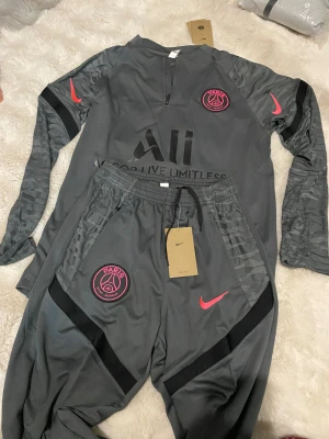 Grå Nike PSG träningsset herr S - Säljer ett grått träningsset från Nike Paris Saint-Germain med långärmad tröja och matchande byxor. Setet har svarta och rosa detaljer, PSG-logga på bröst och ben, samt Nike Swoosh. Materialet är lätt polyester med mönstrade ärmar och dragkedja i halsen.