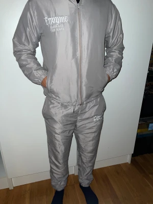 Envyme Full Set - Säljer ett par grå mjukisbyxor från Envyme med broderad logga och texten 'Embrace the Hate' på vänster lår. Byxorna har elastisk midja med snörning och sidofickor. Materialet är glansigt syntet och passformen är loose med smalare benslut. Båda är i storlek S, insidan är varm. Knappast använd inga defekter.